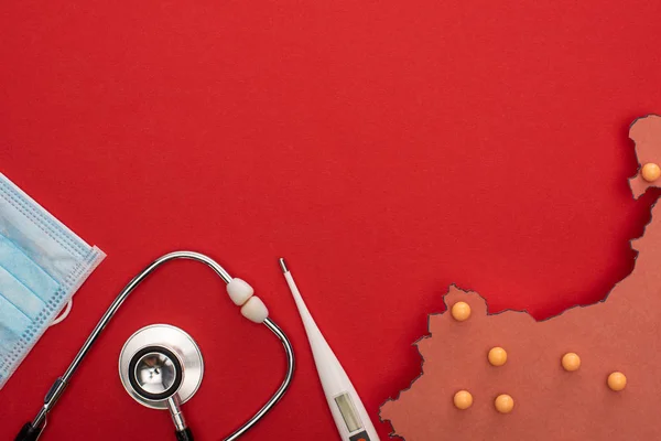 Vista dall'alto di termometro, stetoscopio, maschera medica e layout della mappa della porcellana con spilli gialli su sfondo rosso — Foto stock