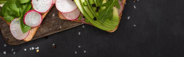 Panoramic shot of healthy sandwiches with fresh radish and avocado on wooden board with pepper and salt — Stock Photo