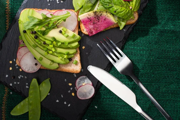 Vue de dessus des toasts végétariens avec des légumes frais sur tissu et planche de pierre avec fourchette et couteau — Photo de stock