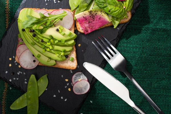 Top view of vegetarian toasts with fresh vegetables on cloth and stone board with fork and knife — Stock Photo