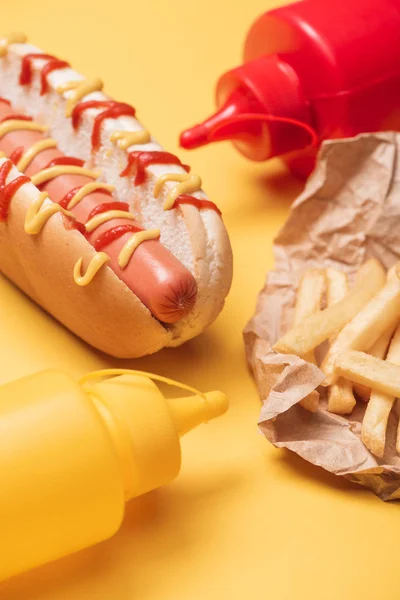 Cachorro-quente, batatas fritas em papel e garrafas com ketchup e mostarda em amarelo — Fotografia de Stock