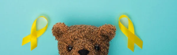 Vue de dessus des rubans jaunes de sensibilisation et de l'ours en peluche sur le concept bleu, panoramique, journée internationale contre le cancer chez les enfants — Photo de stock