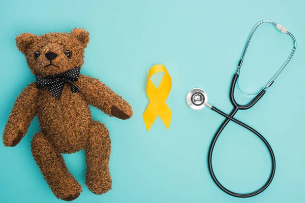 Vista superior de la cinta amarilla de la conciencia, oso de peluche cerca del estetoscopio sobre fondo azul, concepto internacional del día del cáncer de la niñez - foto de stock