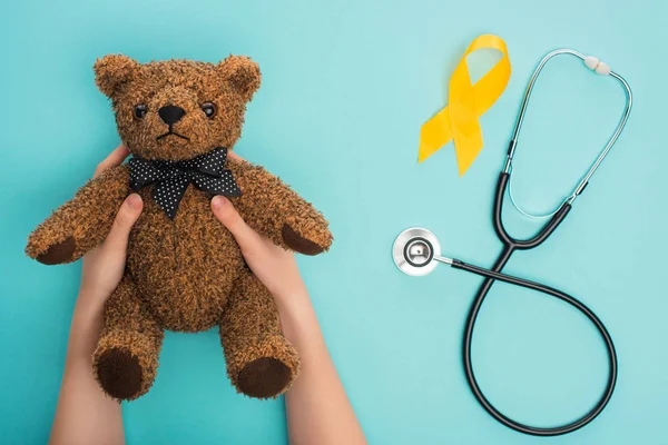 Vista recortada de la mujer sosteniendo oso de peluche cerca de cinta de conciencia amarilla y estetoscopio sobre fondo azul, concepto de día del cáncer infantil internacional - foto de stock