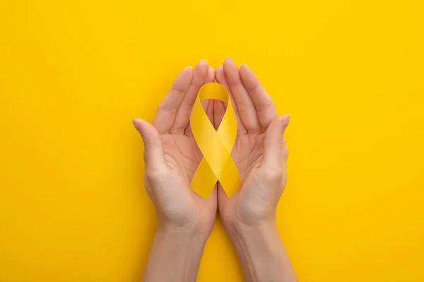 Vue partielle des mains féminines avec ruban de sensibilisation jaune sur fond coloré, concept de journée internationale contre le cancer chez l'enfant — Photo de stock