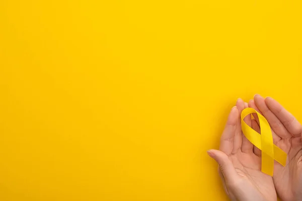 Vista ritagliata di mani femminili con nastro di consapevolezza gialla su sfondo colorato, concetto di giornata internazionale del cancro infantile — Foto stock