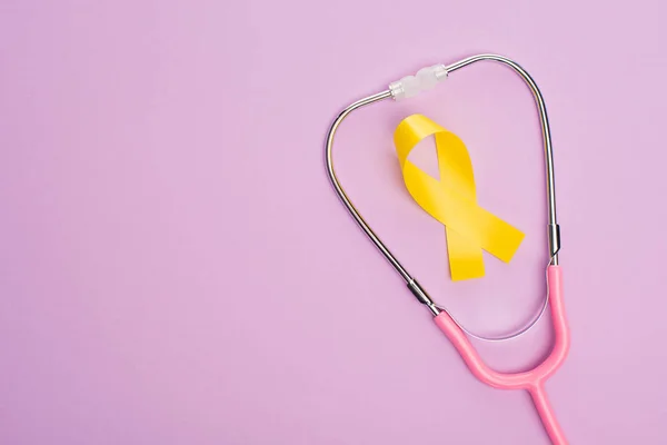 Top view of yellow ribbon and pink stethoscope on violet background, international childhood cancer day concept — Stock Photo
