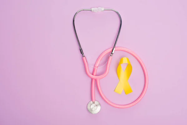 Vista superior de la cinta amarilla con estetoscopio rosa sobre fondo violeta, concepto del día internacional del cáncer infantil - foto de stock