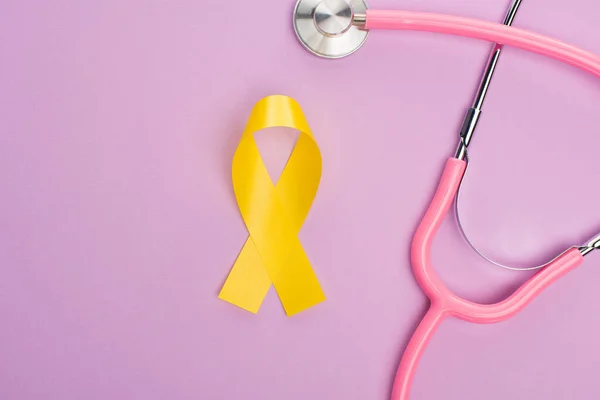 Top view of yellow ribbon and stethoscope on violet background, international childhood cancer day concept — Stock Photo