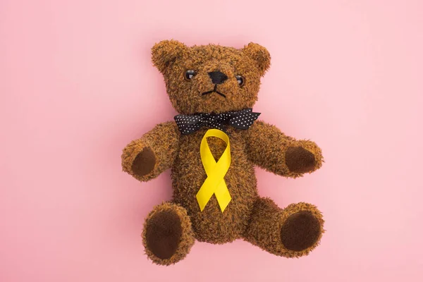 Top view of yellow awareness ribbon on teddy bear with bow on pink background, international childhood cancer day concept — Stock Photo