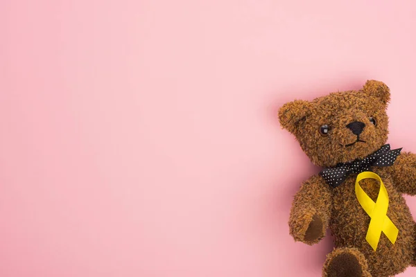 Visão superior da fita amarela no ursinho de pelúcia com arco no fundo rosa, conceito do dia internacional do câncer infantil — Fotografia de Stock