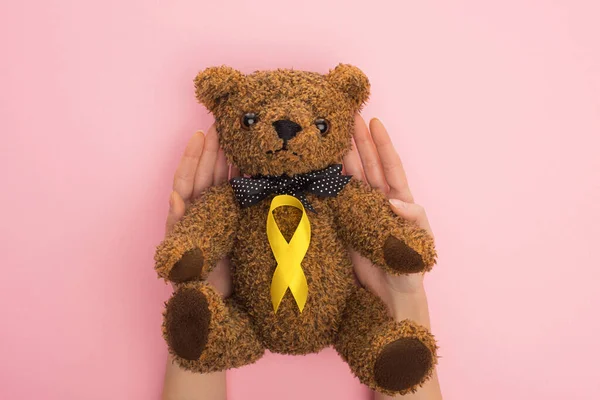 Ausgeschnittene Ansicht einer Frau mit Teddybär mit gelbem Band auf rosa Hintergrund, Konzept zum internationalen Kinderkrebstag — Stockfoto