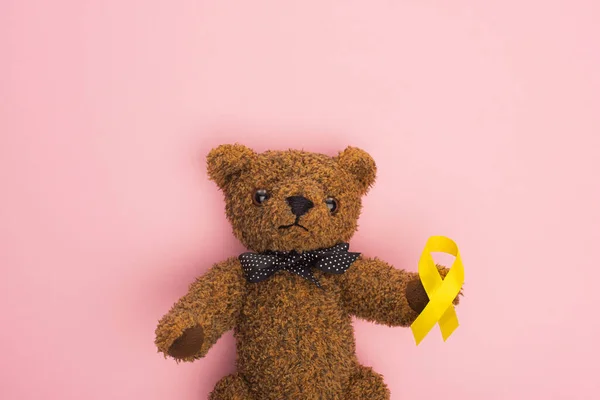 Vista superior da fita amarela no urso de pelúcia marrom com arco no rosa, conceito do dia internacional do câncer infantil — Fotografia de Stock