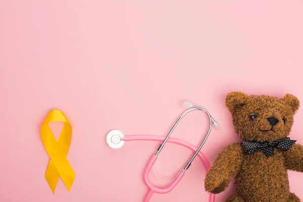 Vista superior de la cinta amarilla cerca del estetoscopio y el oso de peluche sobre fondo rosa, concepto internacional del día del cáncer infantil — Stock Photo