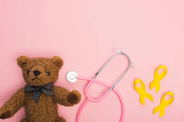 Vue de dessus des rubans jaunes à côté du stéthoscope et de l'ours en peluche sur fond rose, concept de journée internationale contre le cancer chez les enfants — Photo de stock