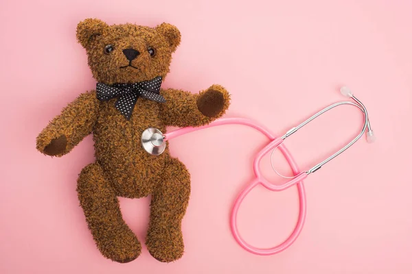 Vista superior del estetoscopio conectado con el oso de peluche sobre fondo rosa, concepto de día internacional del cáncer infantil - foto de stock