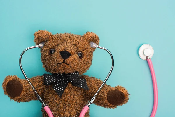 Top view of brown teddy bear with pink stethoscope on blue background, international childhood cancer day concept — Stock Photo