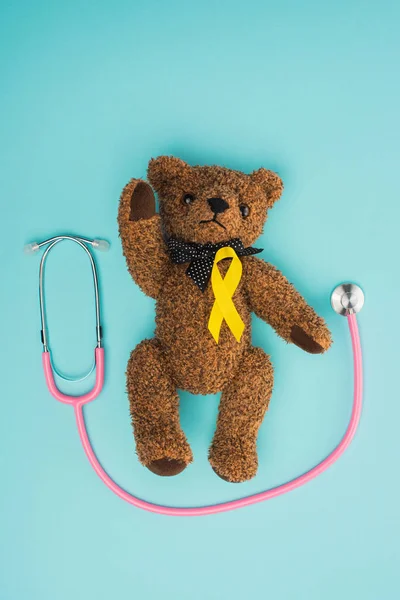 Vista superior da fita amarela no urso de pelúcia marrom perto do estetoscópio no fundo azul, conceito do dia internacional do câncer infantil — Fotografia de Stock