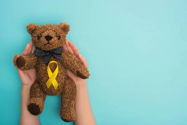 Vista cortada da mulher segurando ursinho de pelúcia com fita amarela no fundo azul, conceito do dia internacional do câncer infantil — Fotografia de Stock