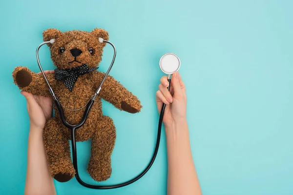 Vista recortada de la mujer sosteniendo oso de peluche con estetoscopio sobre fondo azul, concepto de día internacional del cáncer infantil - foto de stock