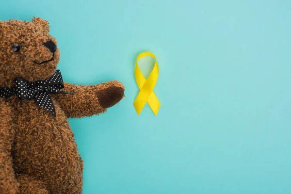 Vue du dessus de l'ours en peluche avec arc et ruban jaune sur fond bleu, concept de journée internationale contre le cancer chez les enfants — Photo de stock