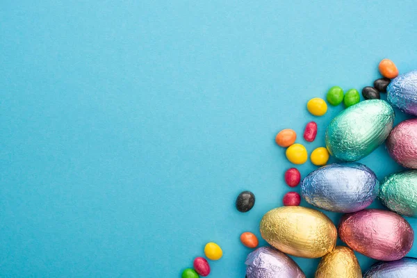 Vue du dessus des œufs de Pâques en chocolat en feuille avec des bonbons colorés sur fond bleu — Photo de stock