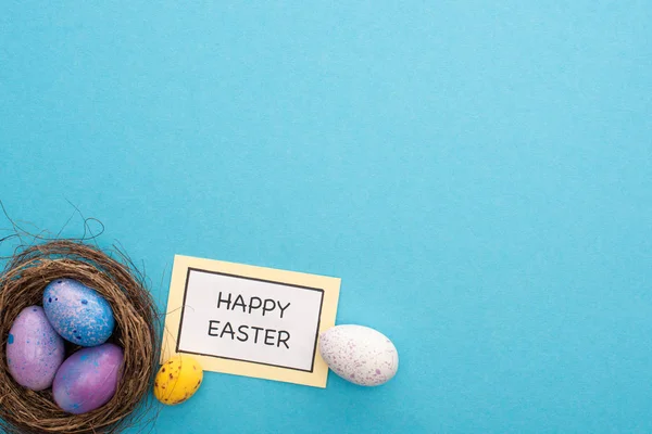 Top view of nest with Easter eggs near card with Happy Easter lettering on blue background — Stock Photo