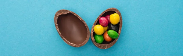 Top view of chocolate Easter egg halves with colorful sweets on blue background, panoramic shot — Stock Photo