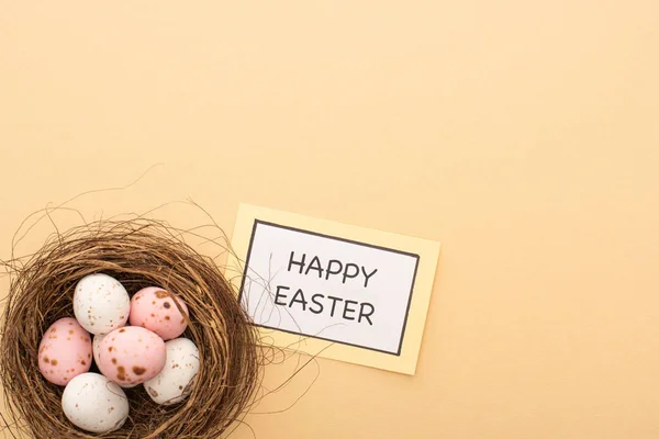 Top view of pink and white quail eggs in nest and card with Happy Easter lettering on beige background — Stock Photo