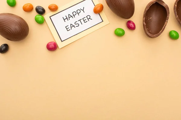 Top view of chocolate eggs, sweets and card with Happy Easter lettering on beige background — Stock Photo