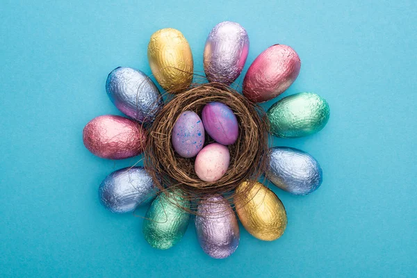 Top view of chocolate Easter eggs in colorful foil around nest with quail eggs on blue background — Stock Photo