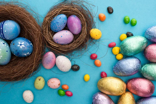 Верхний вид конфет, шоколадные пасхальные яйца в красочной фольге возле гнезд с окрашенной курицей и перепелиными яйцами на синем фоне — стоковое фото