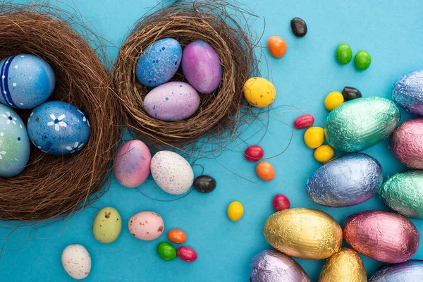 Draufsicht auf Bonbons, Schokoladen-Ostereier in bunter Folie in der Nähe von Nestern mit bemalten Hühner- und Wachteleiern auf blauem Hintergrund — Stockfoto