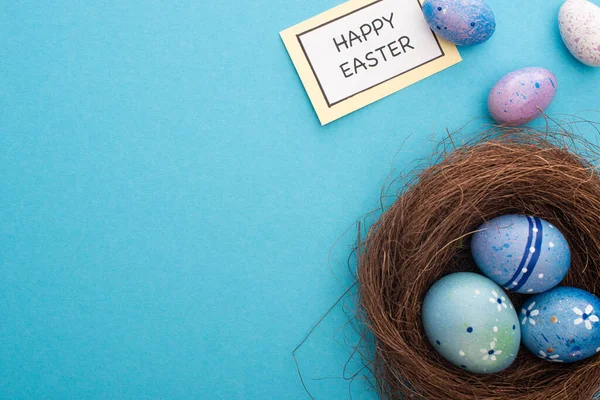 Top view of nest with painted chicken and quail eggs with card with happy easter lettering on blue background — Stock Photo