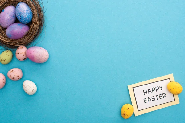 Vista superior del nido con coloridos huevos de pollo y codorniz con tarjeta con letras felices de Pascua sobre fondo azul - foto de stock