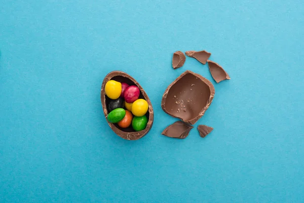 Top view of pieces and half of chocolate Easter egg with colorful candies on blue background — Stock Photo