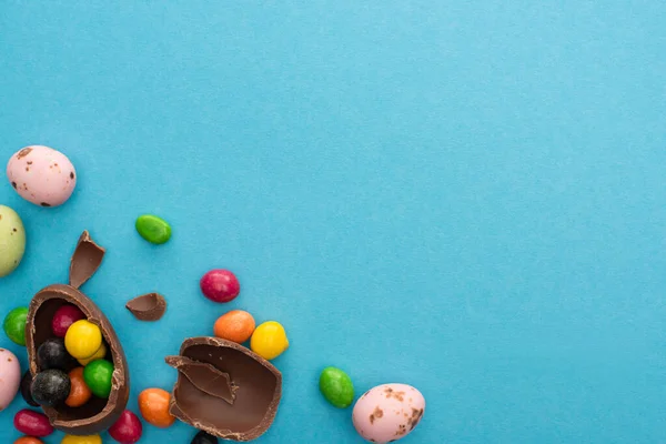 Top view of pieces and half of chocolate Easter egg with candies and colorful quail eggs on blue background — Stock Photo