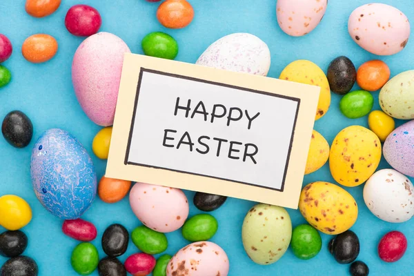 Top view of card with happy easter lettering on colorful chicken and quail eggs, candies on blue background — Stock Photo