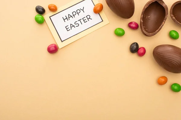 Vista superior de la tarjeta con letras de Pascua feliz con caramelos brillantes y huevos de chocolate sobre fondo beige - foto de stock