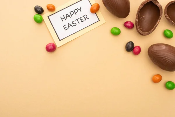 Top view of card with happy easter lettering with bright candies and chocolate eggs on beige background — Stock Photo