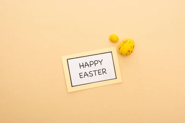 Top view of card with happy easter lettering, candy and yellow quail egg on beige background — Stock Photo