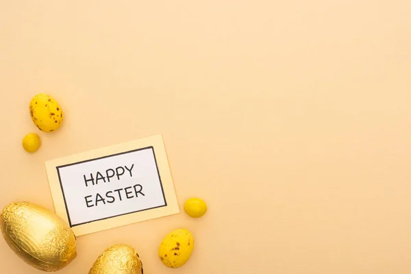 Top view of card with happy easter lettering, candies, yellow quail and chocolate eggs on beige background — Stock Photo