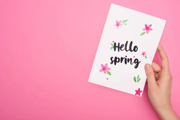 Vue partielle de la carte avec lettrage bonjour printemps dans la main de la femme sur fond rose — Photo de stock