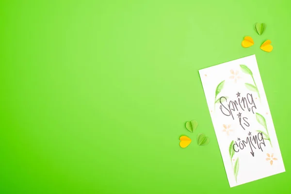 Vista superior de la tarjeta con la primavera está llegando letras y corazones decorativos de colores sobre fondo verde - foto de stock