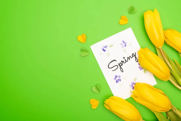 Top view of yellow tulips, card with spring lettering and decorative hearts on green background — Stock Photo