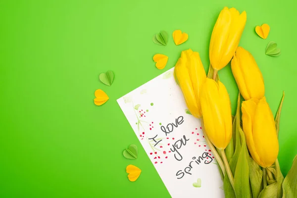 Top view of yellow tulips, card with I love you spring lettering and decorative hearts on green background — Stock Photo