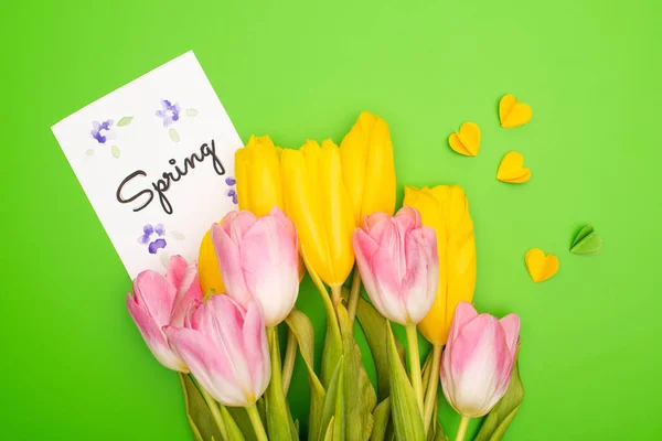 Top view of yellow and pink tulips, card with spring lettering and decorative hearts on green background — Stock Photo