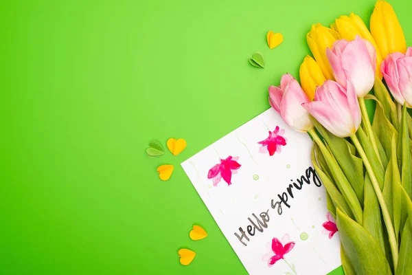 Vista superior de tulipanes amarillos y rosados, tarjeta con letras hola primavera y corazones decorativos sobre fondo verde - foto de stock