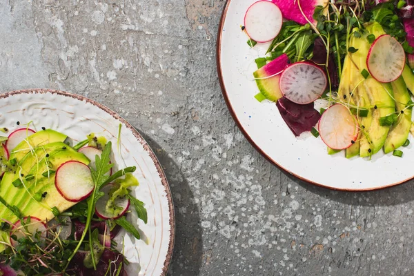 Vista dall'alto di insalata di ravanello fresco con verdure e avocado servito su piatti su superficie di cemento grigio — Foto stock