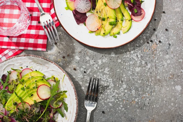 Top view of fresh radish salad with greens and avocado served on plates on grey concrete surface with water, cutlery and napkins — Stock Photo